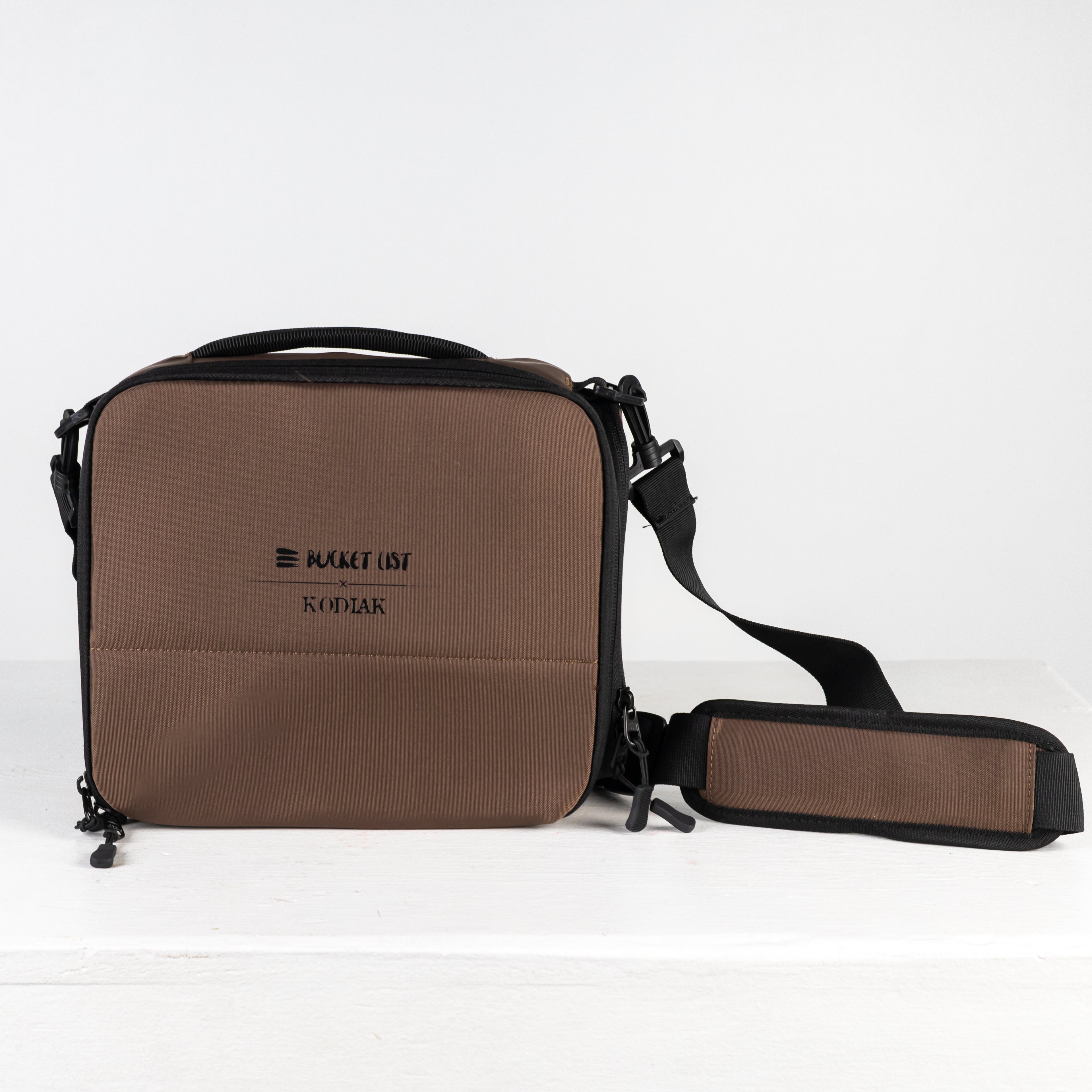 Product photo of a brown camera cube bag on a white table with a white background. The bag has the words Bucket List and Kodiak printed on it in small black letters.