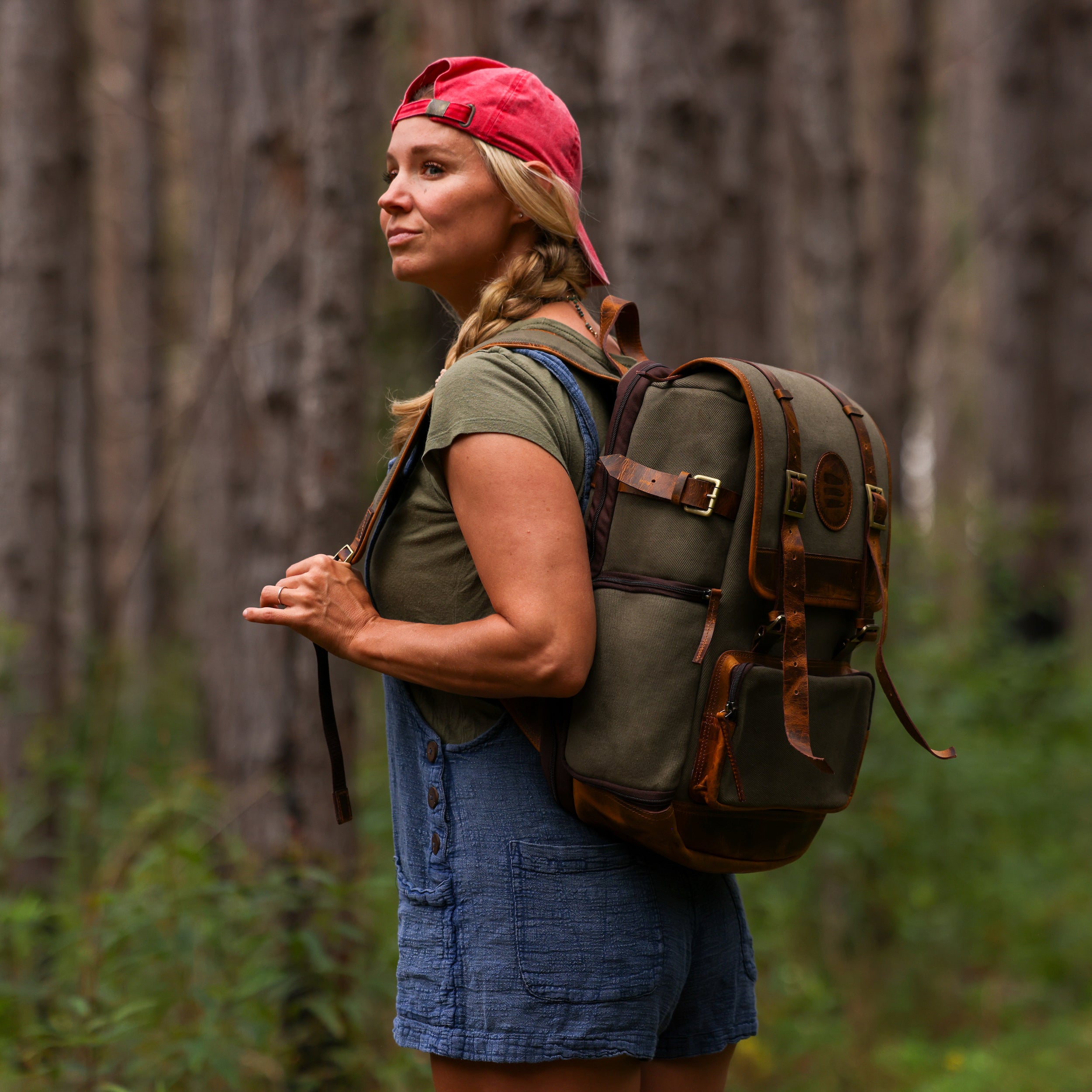 Photo of woman wearing a red baseball cap and a green canvas and leather backpack in the woods.