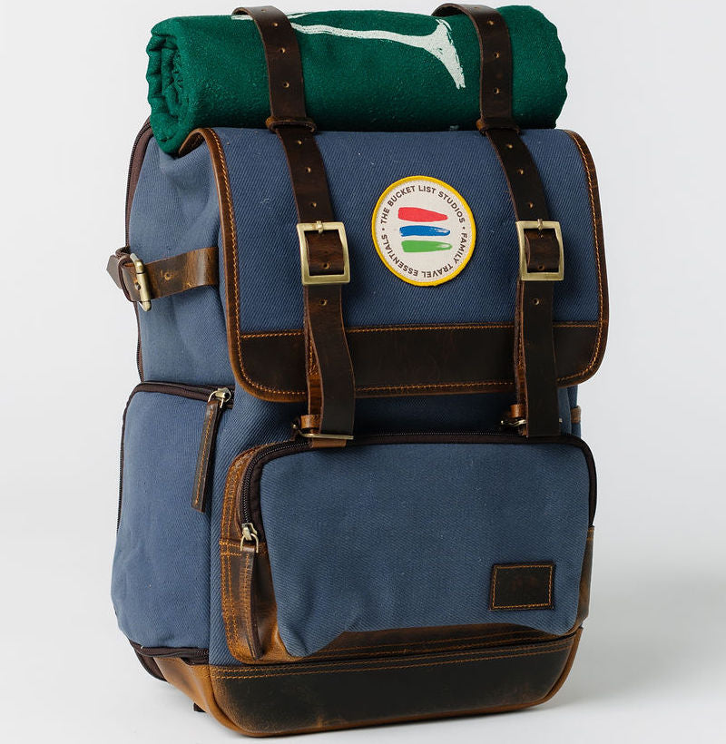 Product photo of a navy canvas and leather backpack with a green rolltop blanket on top of the bag.