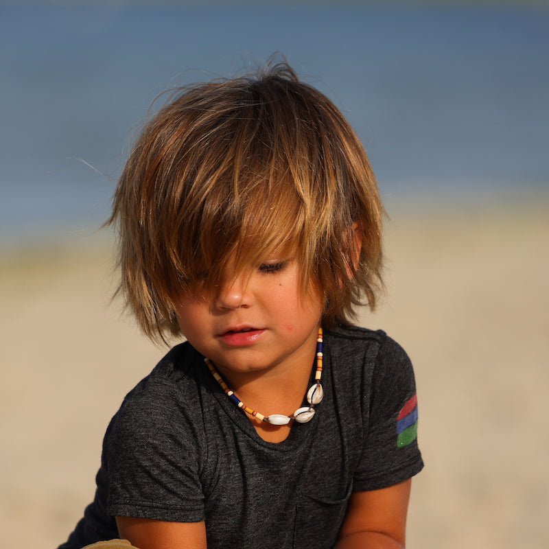 Photo of a young boy on the beach wearing a necklace that has some natural beads and three shells.