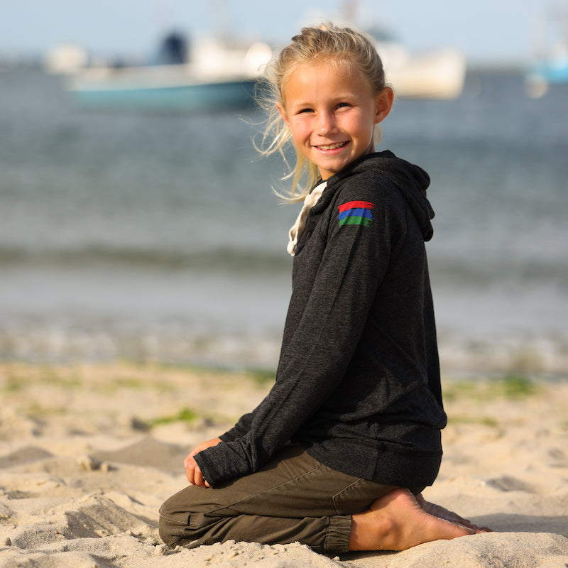 Photo of a girl sitting on her knees on the beach wearing a black hoodie with a red, blue, and green bucket list logo on her arm.