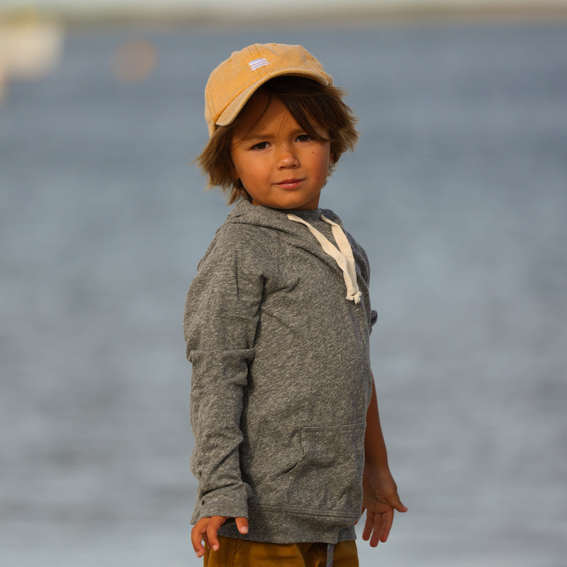 Photo of a young boy in front of the ocean wearing a grey hoodie and a yellow baseball cap.