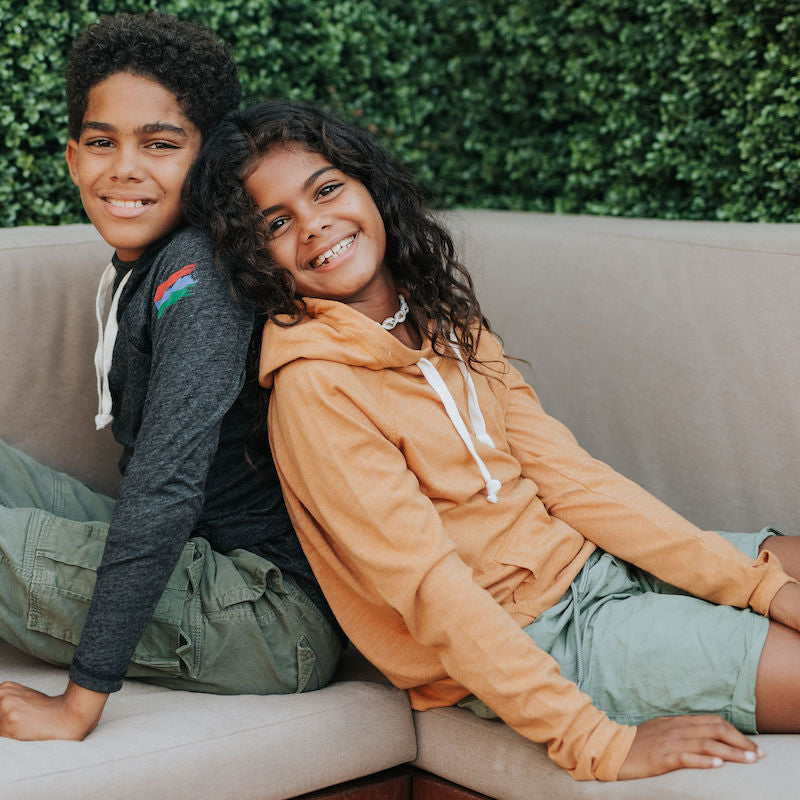 Photo of a brother and sister in a black and a mustard hoodie. They're sitting back to back on an outdoor couch.