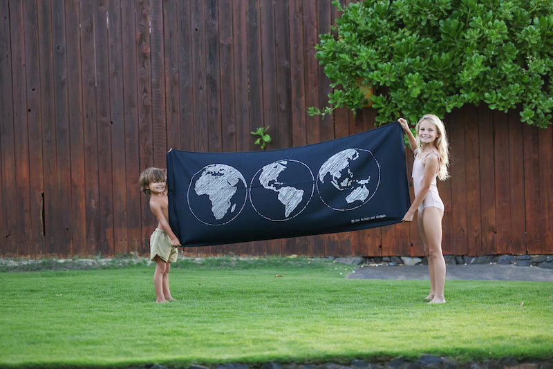 Two kids in a yard holding up a long blanket. The blanket is black with three white globes on it.