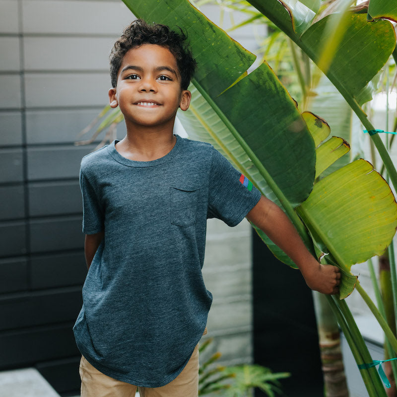 Photo of a boy standing by a large plant wearing a navy short sleeve crew-neck t-shirt with a front pocket and the bucket list logo on his right arm.
