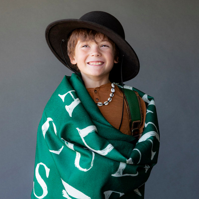 Studio photo of a young boy wearing a brown round brim hat with a leather strap and a green blanket wrapped around his shoulders.
