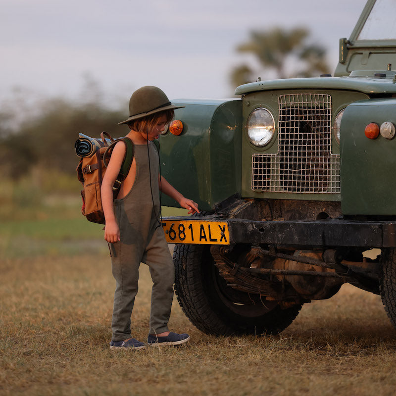 Photo of a boy in front of a green safari car wearing olive overalls, an olive brimmed hat, and a canvas and leather backpack.