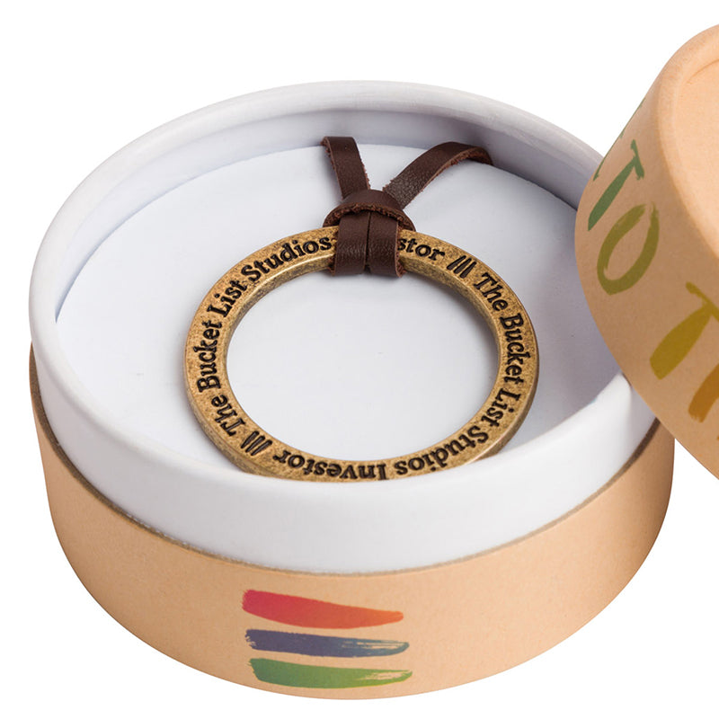 Product photo of a necklace inside of a brown circle necklace box. The necklace inside has a brown leather chain with a circle metal medallion as the focal point of the necklace.