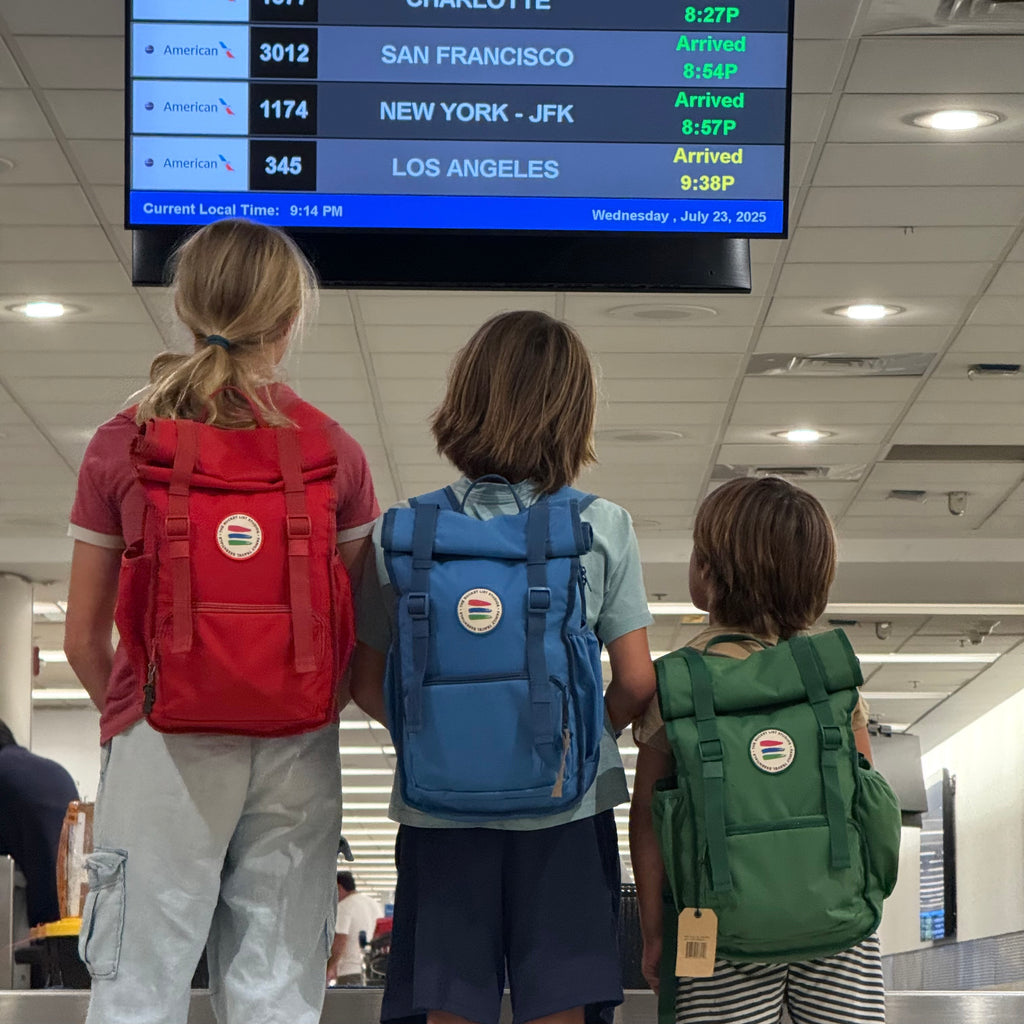 Three children with backpacks looking at an airport arrival screen.