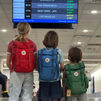 Three children with backpacks looking at an airport arrival screen.