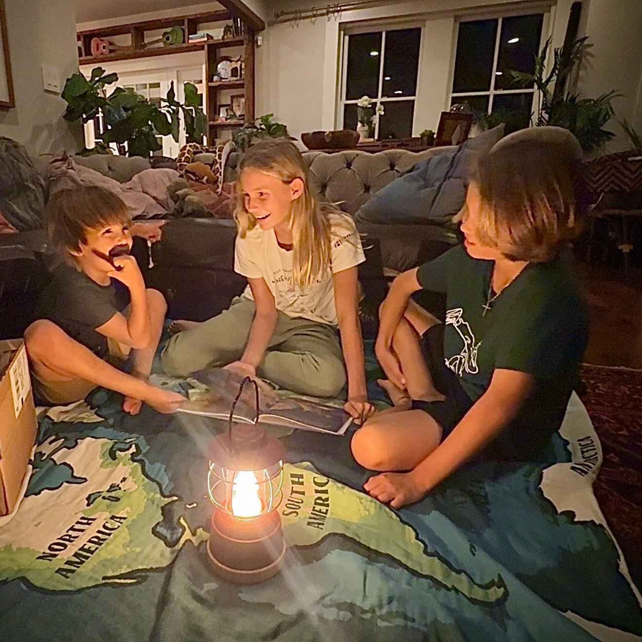 Three children sitting on a blanket with a map, using a lantern, in a cozy living room.