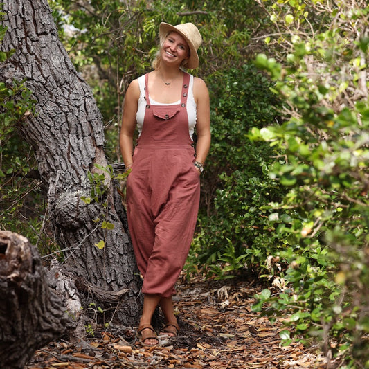 Photo of a woman in the woods wearing mauve overalls with a white tank underneath. 