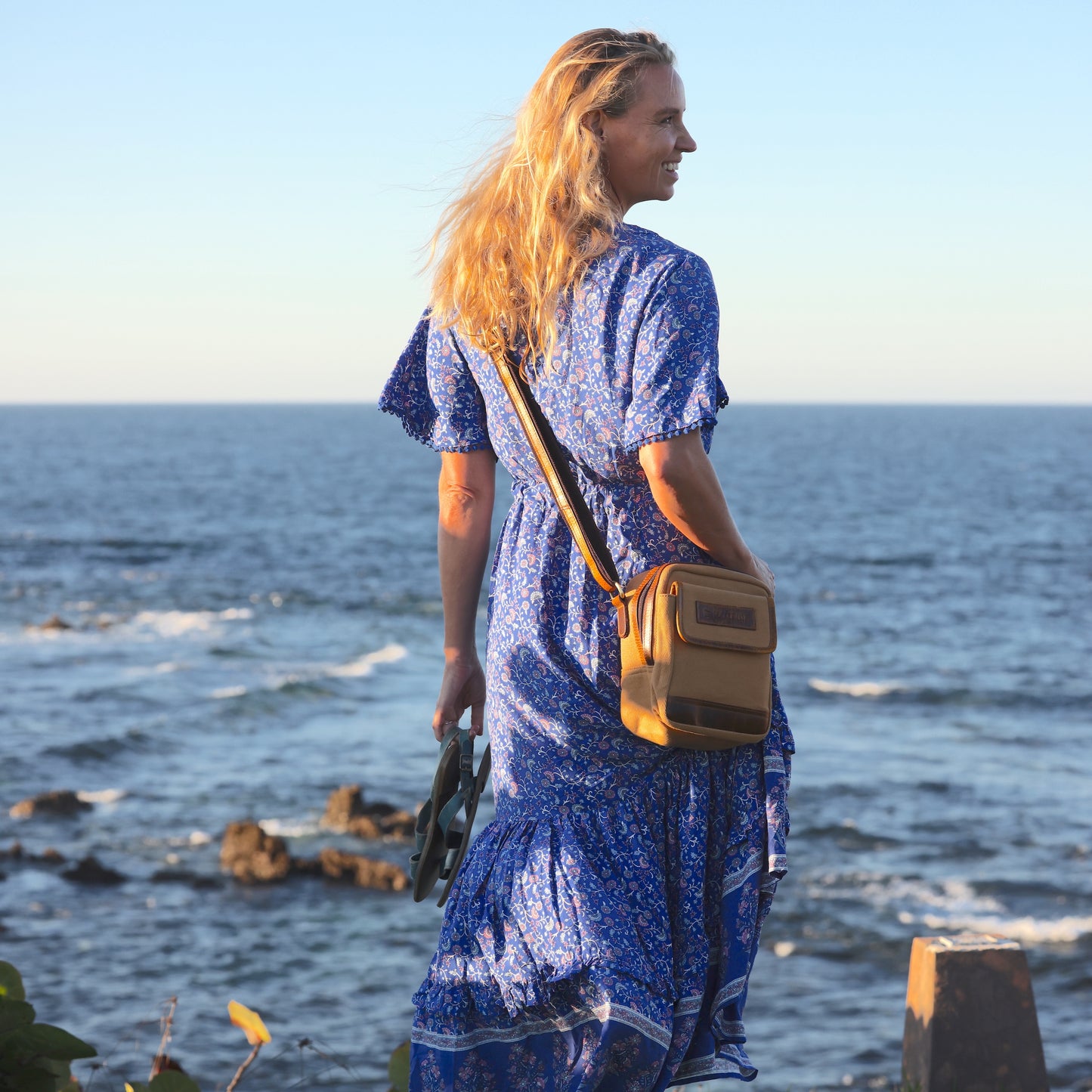 Photo of a woman wearing a dress and a tan sling pouch on the coast. 