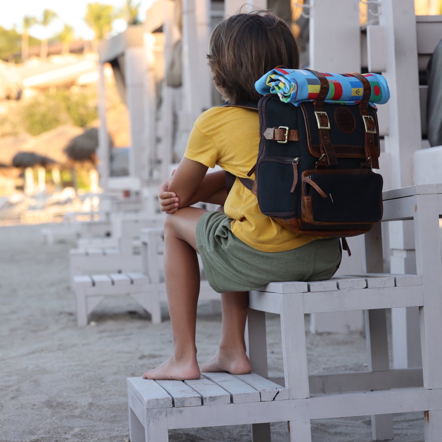 Photo of the back of a  boy sitting on the steps of a lifeguard tower wearing a black canvas and leather backpack.