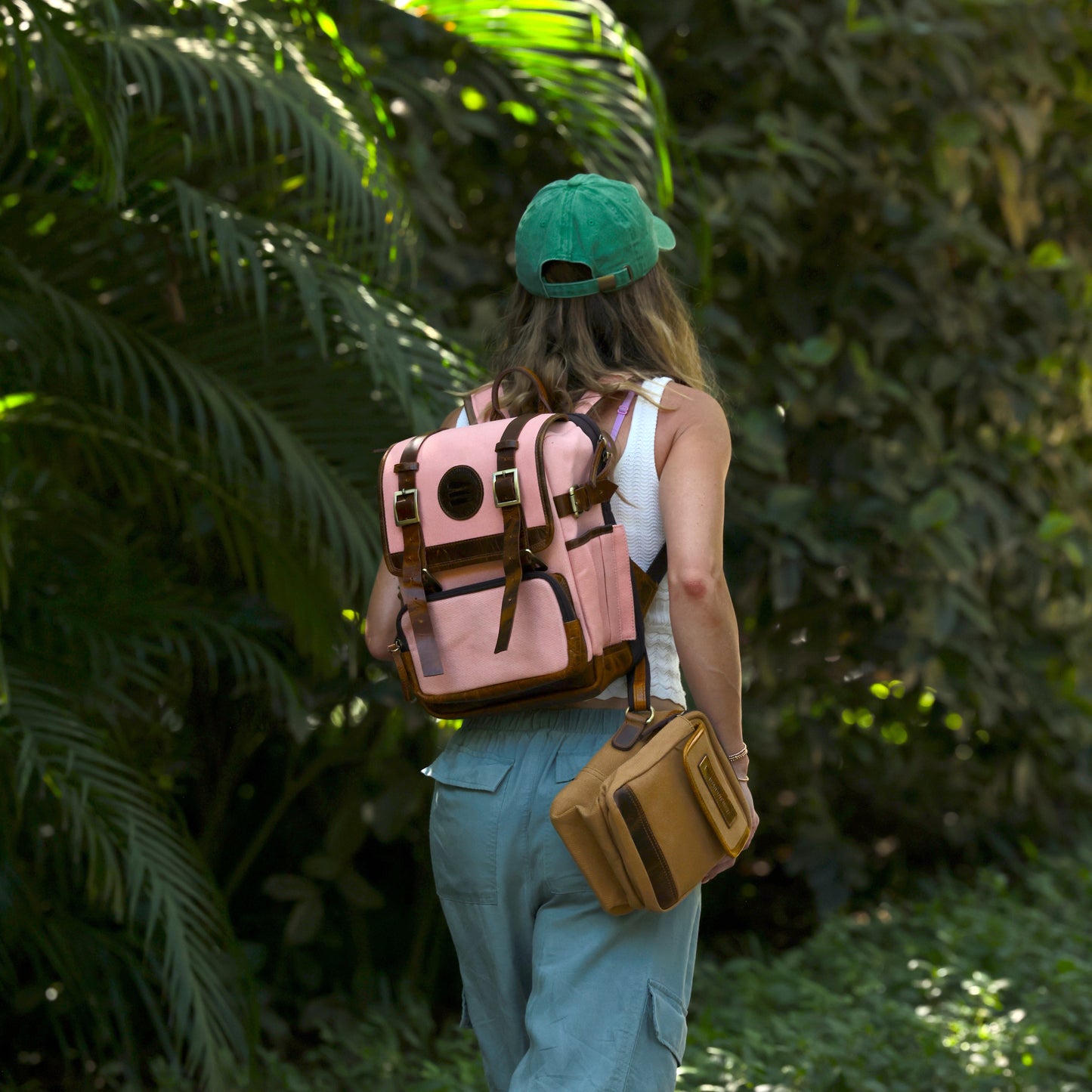 Photo of a woman walking away wearing a pink canvas and leather backpack. She is also wearing a green cap and tan side bag.