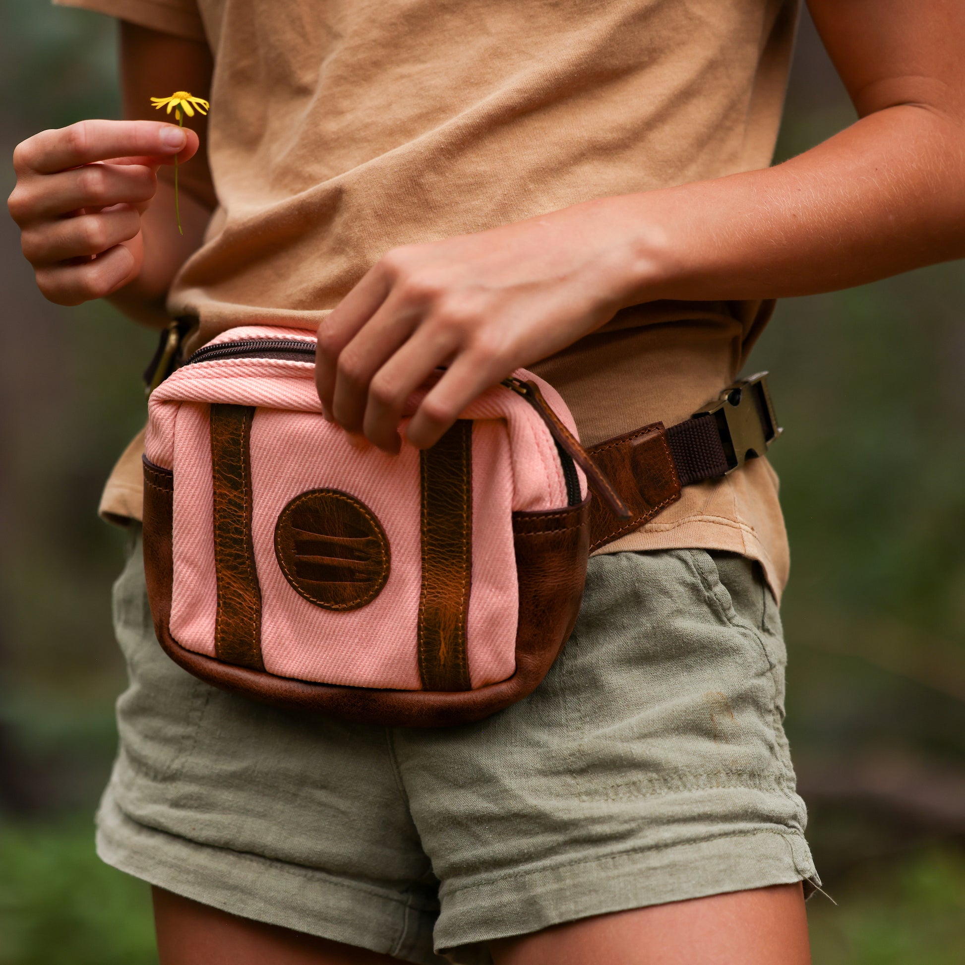 Photo of a girl wearing a pink canvas and leather fanny pack around her waist while holding a small yellow flower.