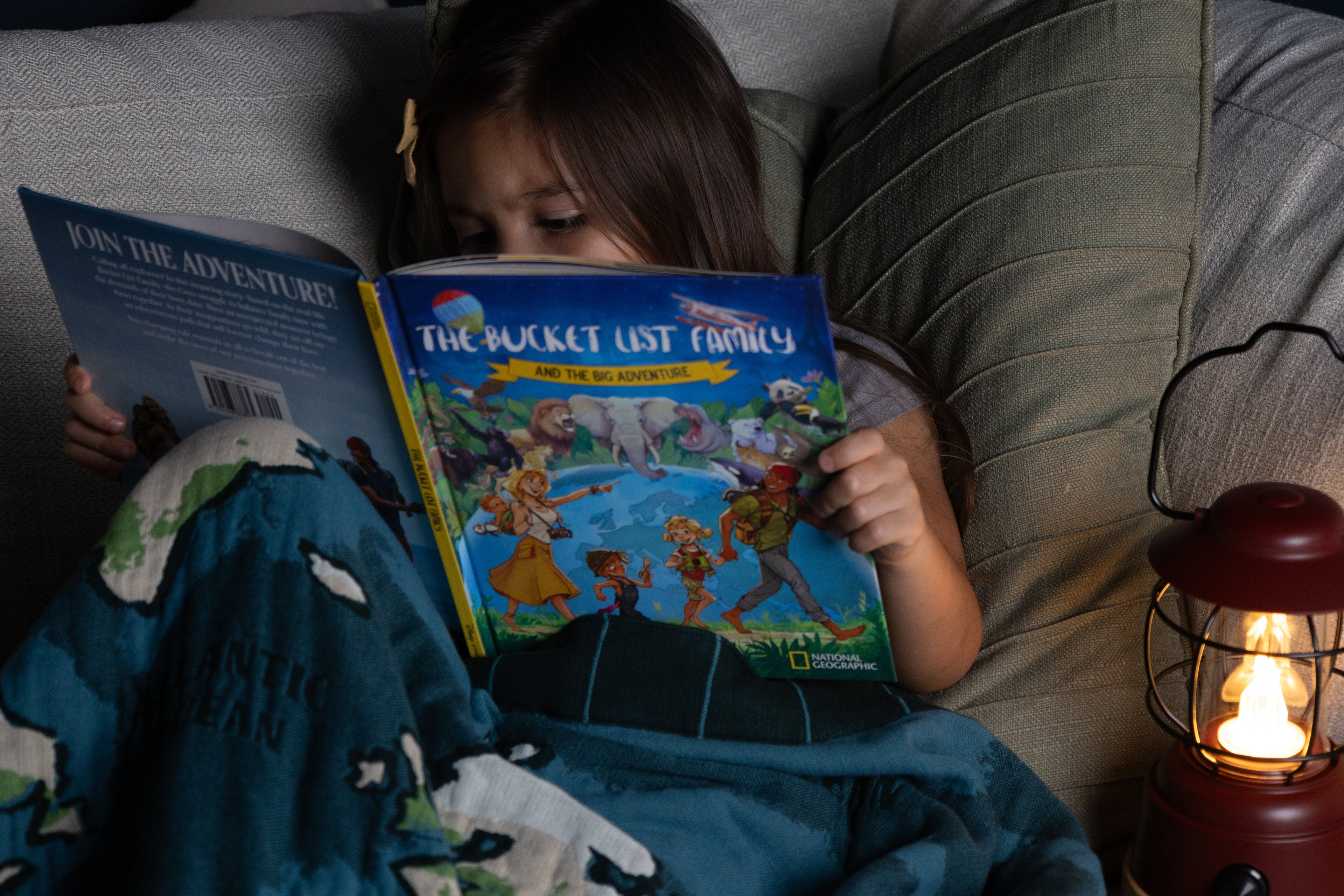 Child reading a book titled 'The Bucket List Family' on a couch with a warm lantern light source.