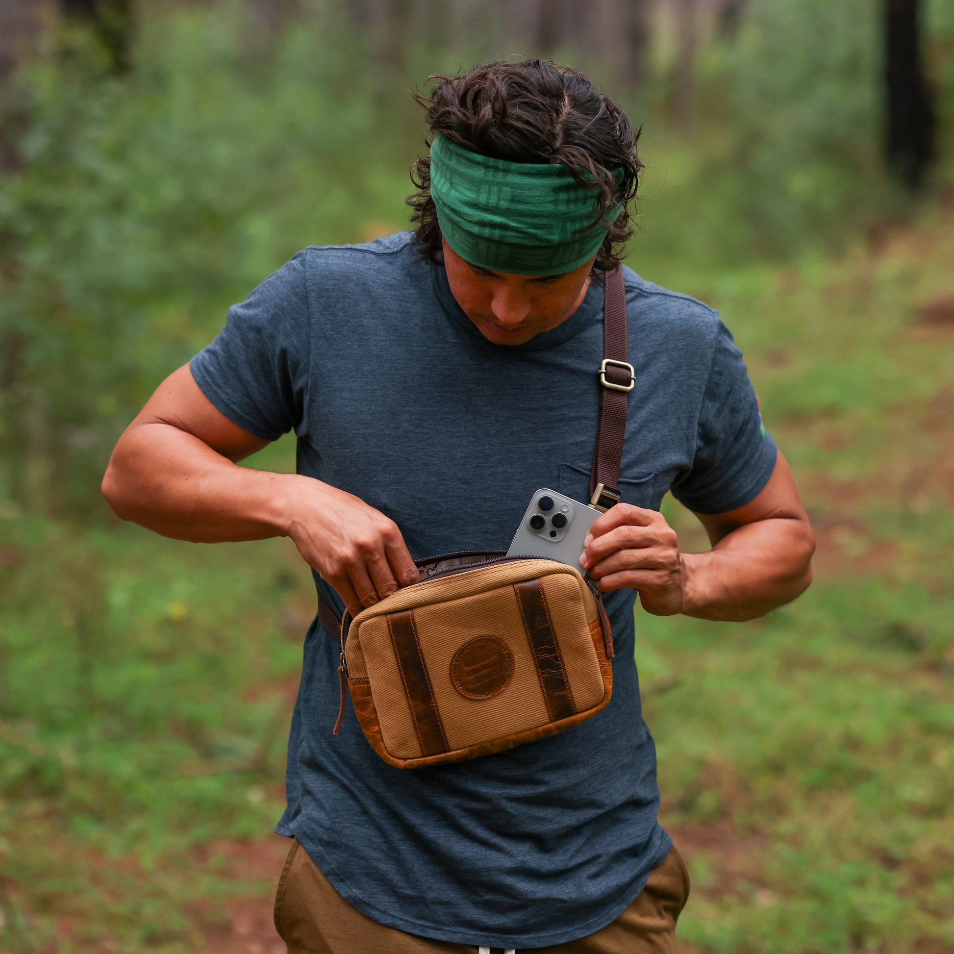 Photo of a man in the woods wearing a tan canvas and leather crossbody belt bag across his chest and putting his iPhone into the bag.