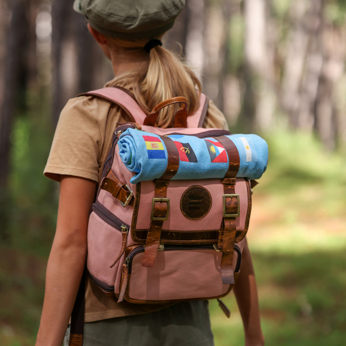 Image of the back of a girl wearing a pink canvas and leather backpack. There is a light blue blanket strapped to the top of the backpack. 