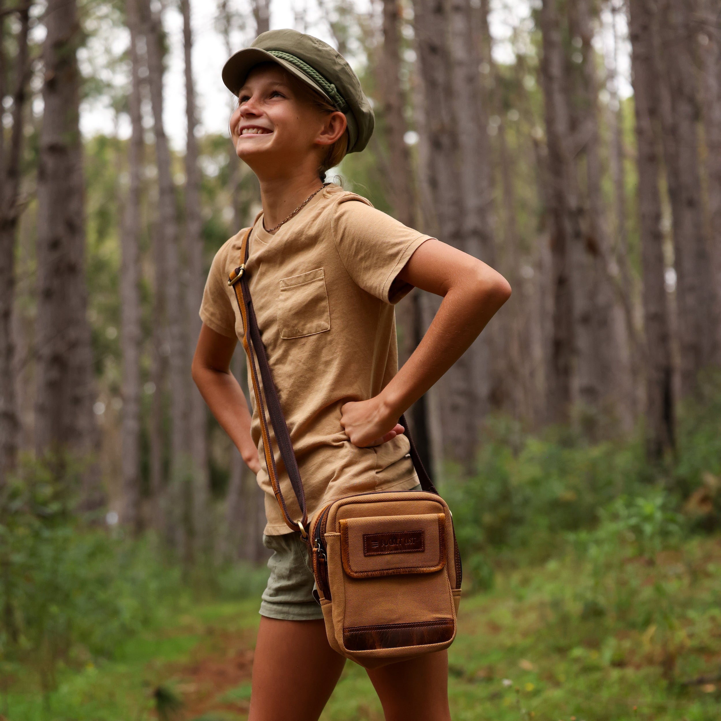 Photo of a girl standing in the woods with her hands on her hips with a tan canvas and leather crossbody bag by her hip. 