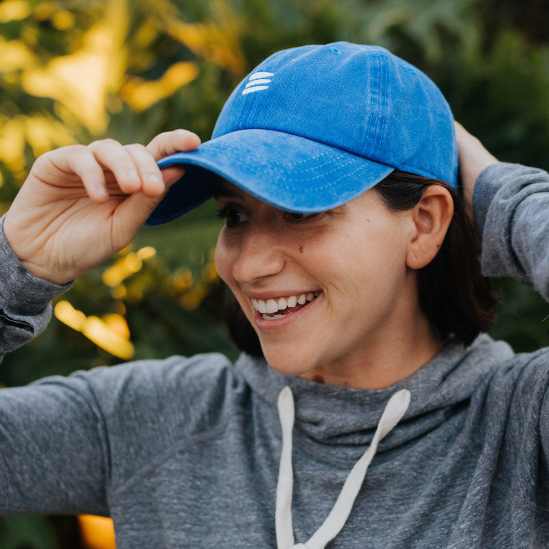 Photo of a woman wearing a grey hoodie and a blue baseball cap with a white bucket list logo embroidered on the front. 