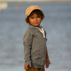 Photo of a young boy in front of the ocean wearing a grey hoodie and a yellow baseball cap.