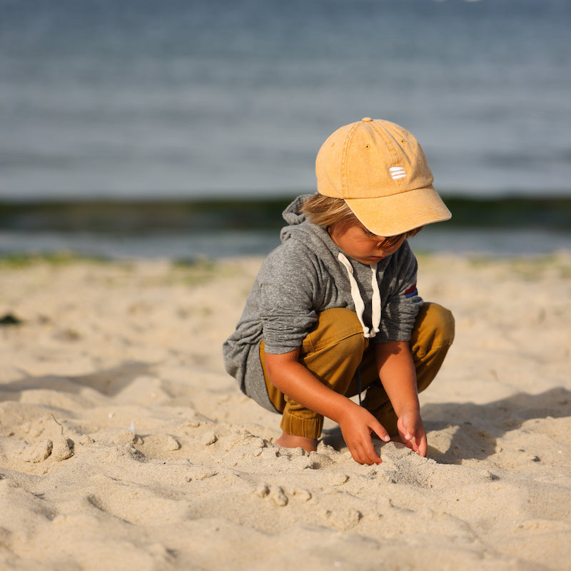 Photo of a little boy on a beach wearing a grey hoodie with a yellow baseball cap that has a white bucket list logo embroidered on the front of the cap.