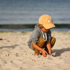 Photo of a little boy on a beach wearing a grey hoodie with a yellow baseball cap that has a white bucket list logo embroidered on the front of the cap.