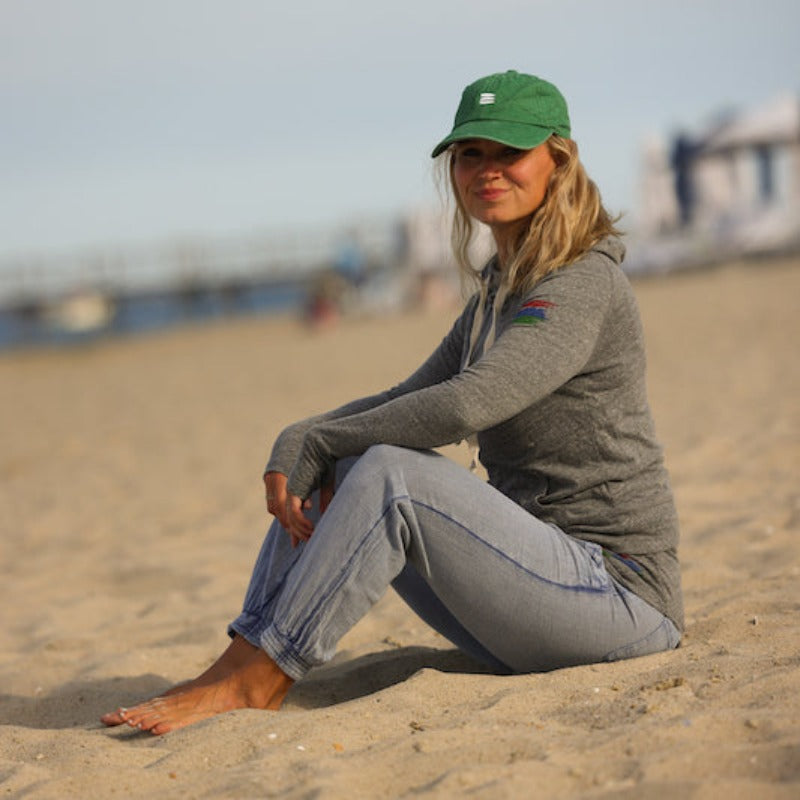 Photo of a woman sitting on a beach looking at the camera. She is wearing a grey hoodie and a green baseball cap with a white bucket list logo embroidered on the front. 