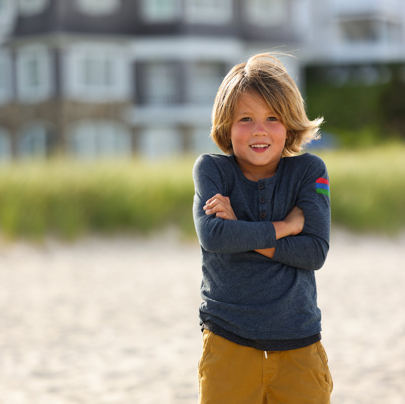 Photo of a young boy standing on a beach wearing a navy henley. 